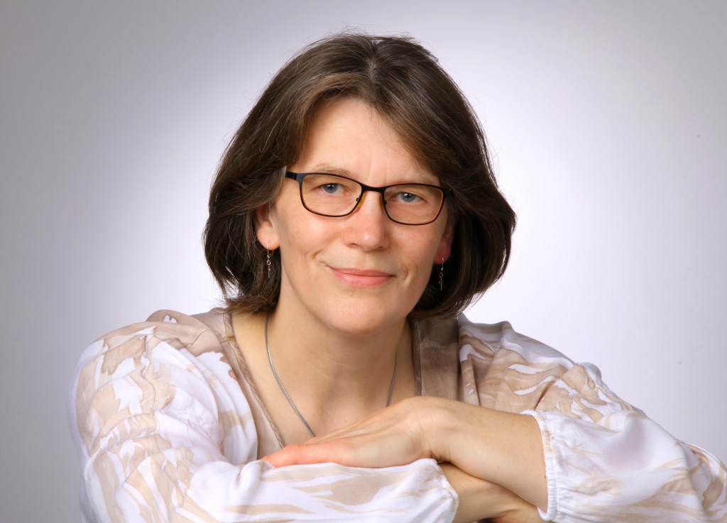 Porträt einer lächelnden Frau mit kurzen, braunen Haaren und Brille vor einem neutralen, hellgrauen Hintergrund. Sie trägt ein gemustertes Oberteil in Weiß- und Beigetönen, eine dezente Halskette sowie Ohrringe und hat ihre Arme vor sich verschränkt, auf denen sie sich leicht abstützt.